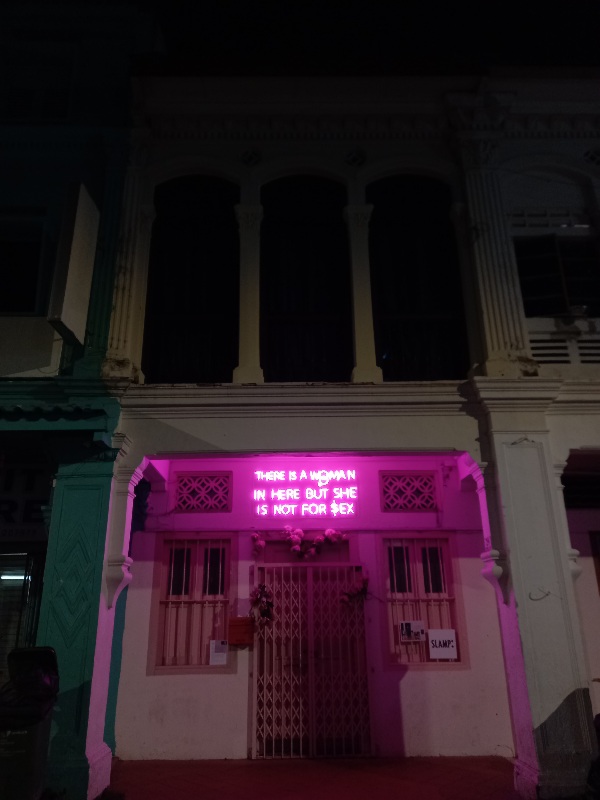 Above a door to a house along Rowell Road,a pink neon sign proclaims "THERE IS A WOMAN IN HERE BUT SHE IS NOT FOR $EX"